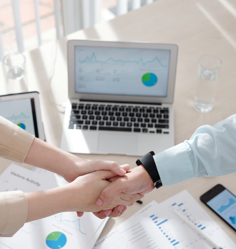 Hands of business people shaking hands over table with various charts and reports after having successful meeting